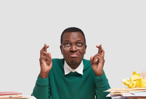Wishful African American man crosses fingers for good luck, believes in fortune dressed in green elegant sweater, surrounded with paper documents, isolated over white background with free space